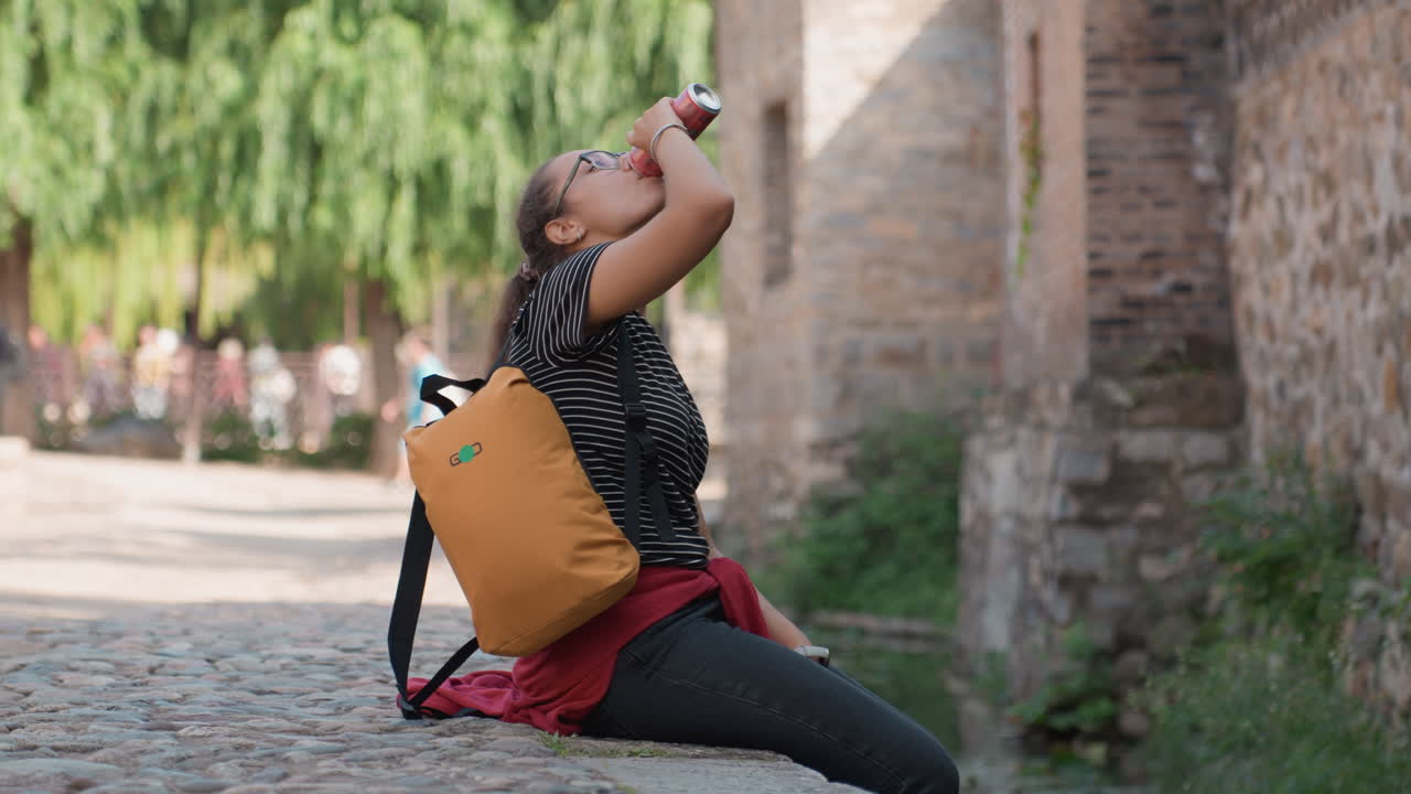 Woman seated on cobblestone bench drinking water. Caucasian woman pauses on rustic bench to hydrate in summer. Traveling woman with orange backpack takes moment to drink water on cobblestone seat
