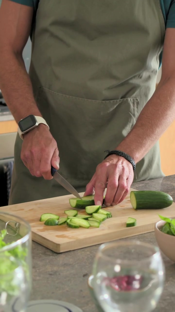 Video verticale: Uomo che afferra un coltello e indossa un grembiule, affettando un cetriolo su un tagliere mentre prepara un'insalata in cucina.