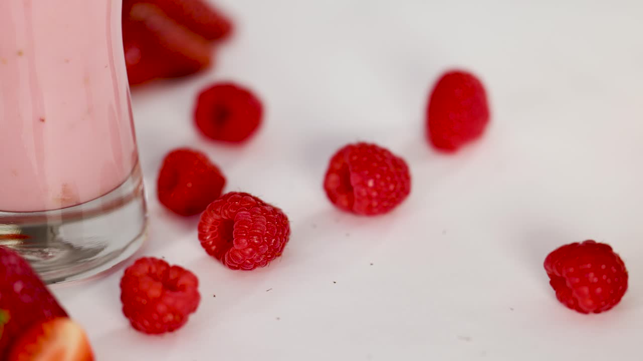 Hand arranges fresh raspberries near pink strawberry milkshake in bright, soft studio lighting