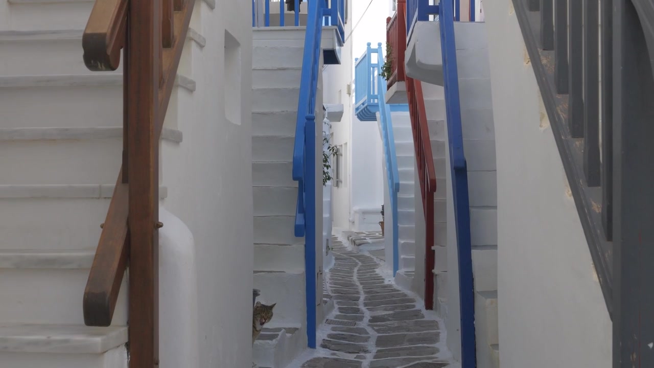 Stretto vicolo acciottolato e tortuoso in un villaggio cicladico con edifici imbiancati a calce e ringhiere colorate. Inquadratura dall'alto verso il basso.