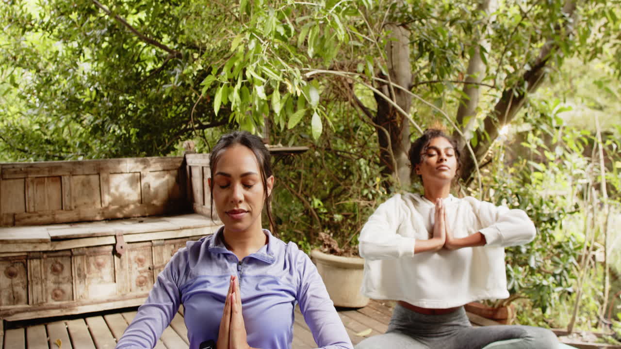 Multiracial female friends meditating on porch, practicing yoga with peaceful expressions in nature
