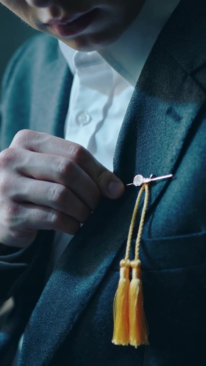 Vertical video: Preparing for event in prep, man fastening lapel pin to dark blazer, gold tassels