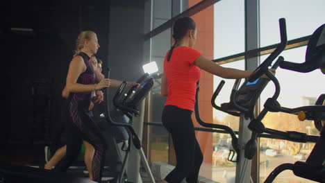 young fit woman using an elliptic trainer in a fitness center. a group of young women train on sports training equipment in a fitness gym. steady cam shot.