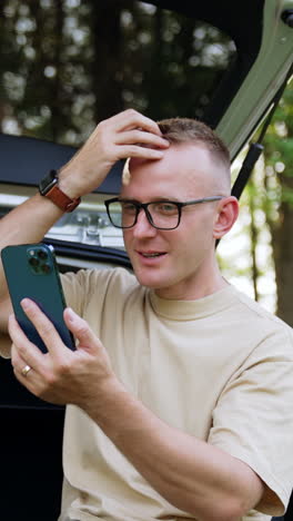 caucasian male in glasses looks at smartphone. man sitting in his car trunk talks on the phone and check his hair on the gadget screen. vertical video