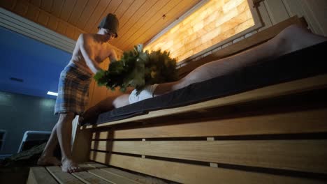 man lying on wooden bench in spa with masseur doing massage with oak broom. client in finn sauna indoors with professional.