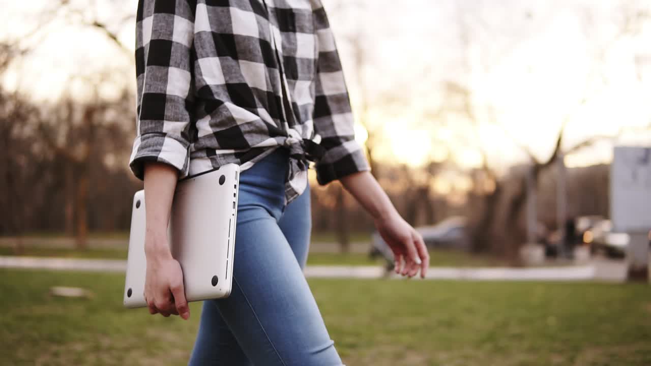 Slim and tender girl in jeans and shirt walking by green side outside, holding laptop in right hand. The light day is running out. Close up. Without face