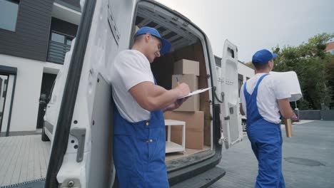 two young workers of removal company unload boxes and furniture from minibus into customer's home