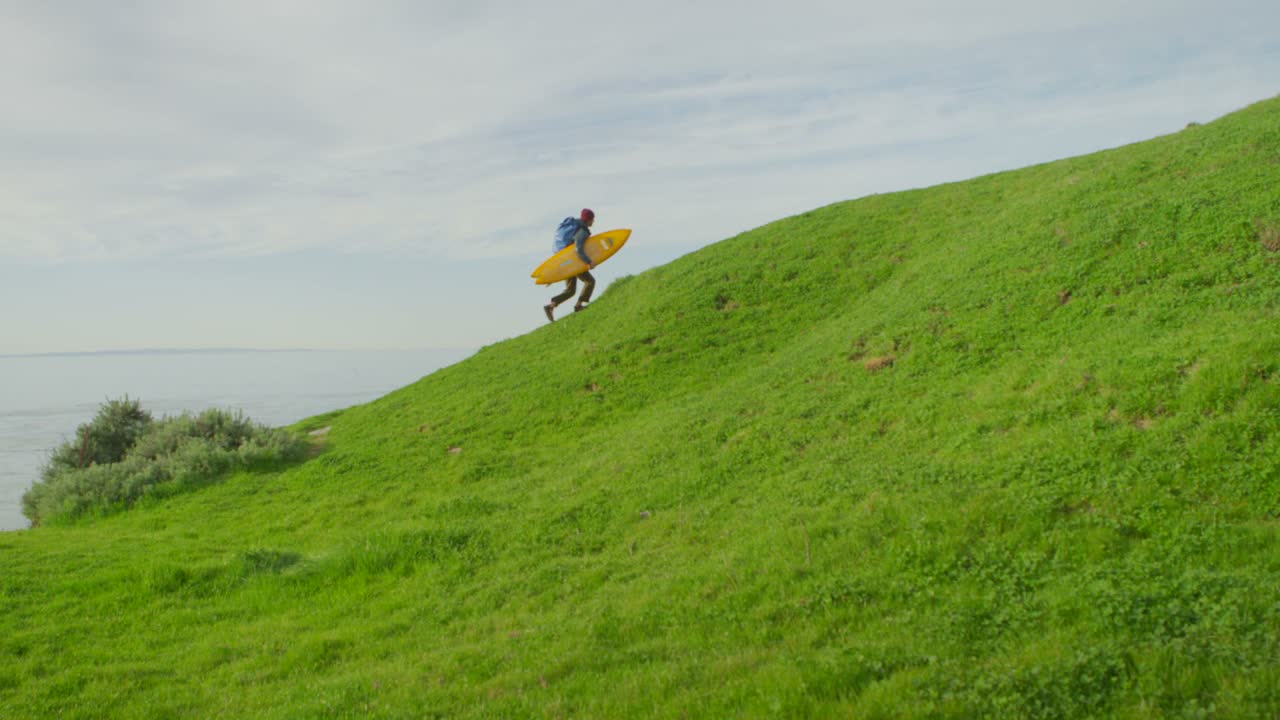 한 서퍼가 해안 지역의 외딴 서핑 장소로 하이킹하면서 보드를 언덕 위로 옮깁니다.