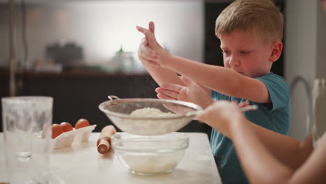 boy gently dusts flour from his hand above sieve while woman sifts flour into bowl on counter, with background showing eggs, rolling pin, and clear glasses during relaxed home baking scene