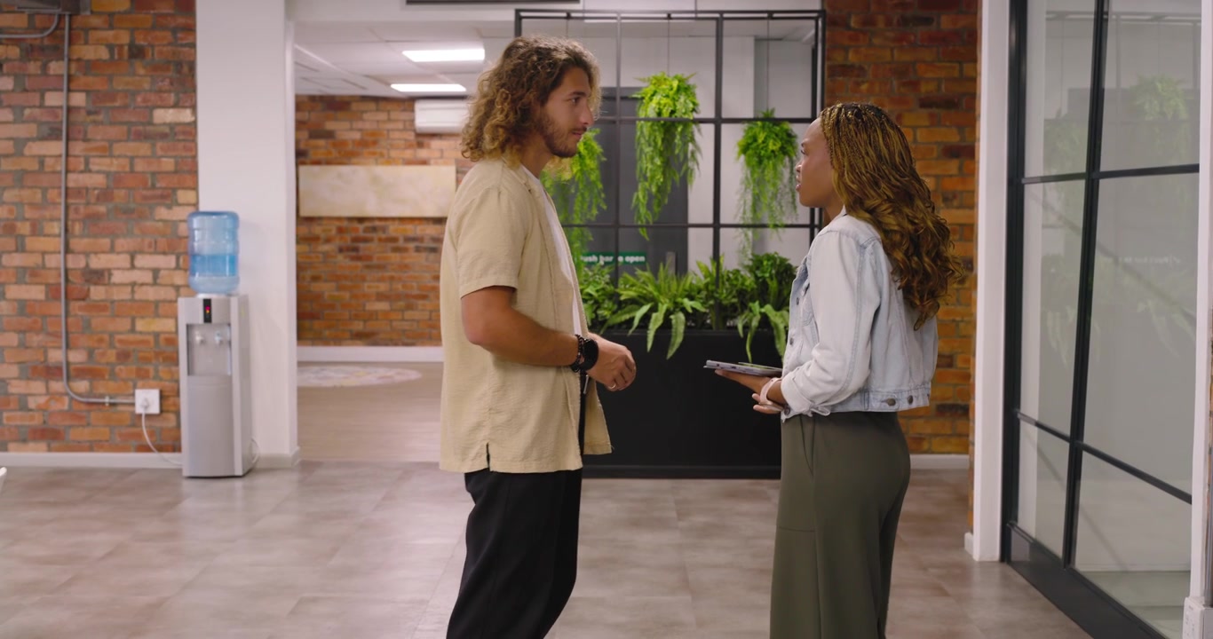 Compañeros de trabajo diversos caminando por el pasillo de la oficina, sosteniendo una tableta, acercándose a una jardinera e iniciando una discusión.