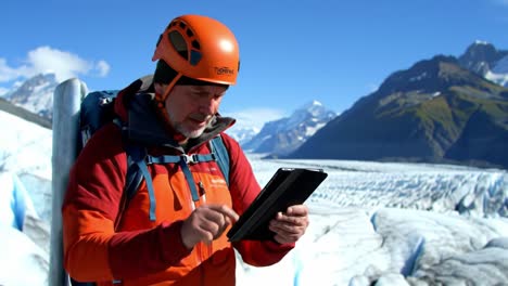 an experienced explorer is engaged with a tablet in a breathtaking mountainous region, surrounded by glaciers and clear blue skies.