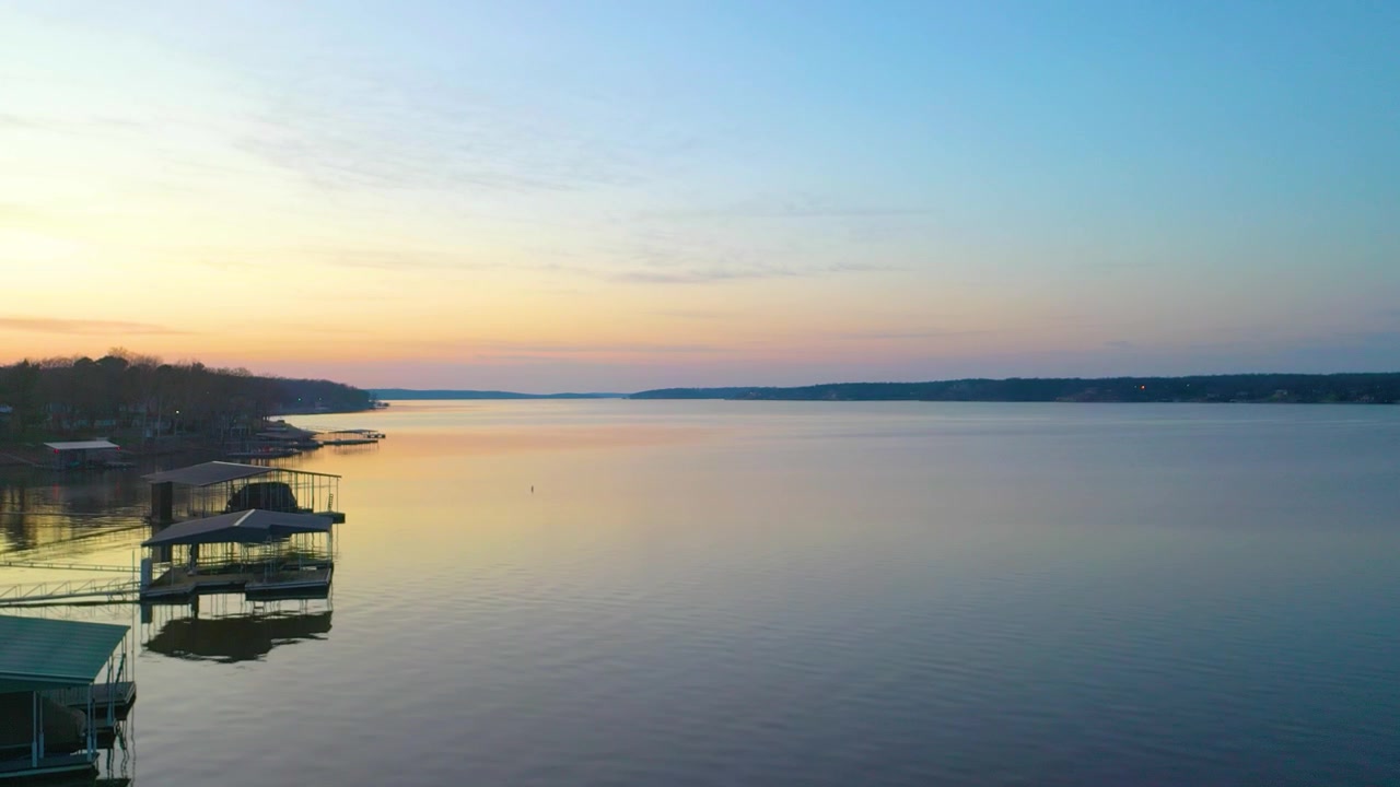 Riprese aeree con drone al tramonto di un pittoresco rifugio sul lago con riva alberata, residenze sul lago, pontili coperti per barche, acqua calma e riflettente e un cielo colorato sopra la baia alla luce della sera