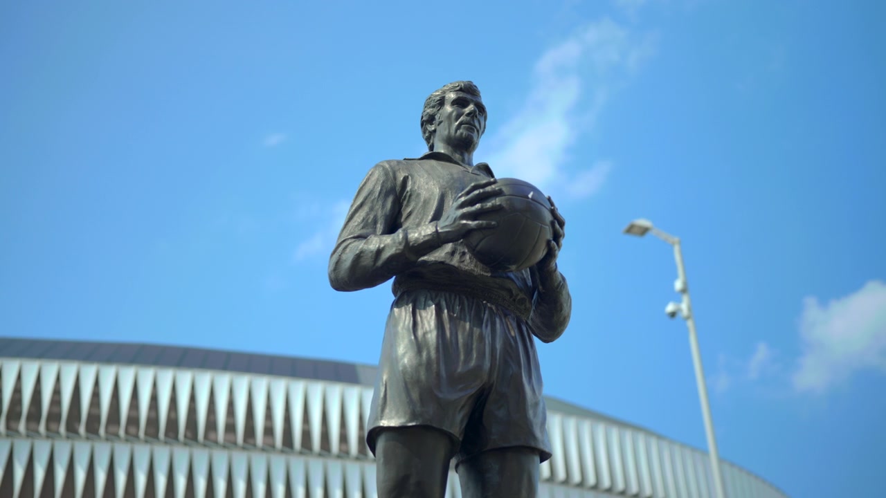 Statue d'athlète en bronze tenant un ballon de cuir vintage au niveau de la poitrine devant un stade moderne, vue d'un point de vue bas avec une patine prononcée, des ombres nettes, un ciel bleu et une présence d'art public monumentale.