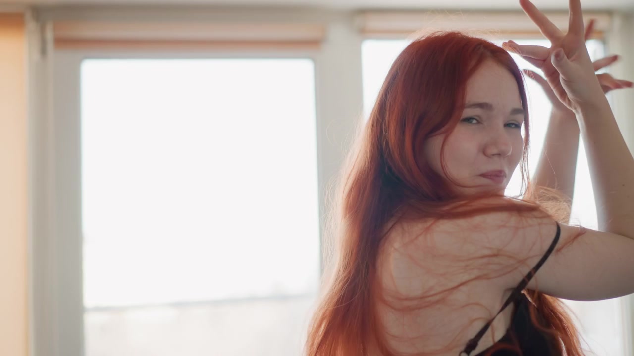 活発なダンサーが光に照らされる。窓際で踊る躍動的な女性の情景。窓のそばで腕を流れるように動かす活気ある女性ダンサーの姿。スタジオの窓辺で楽しそうに踊る女性の親密なショット。