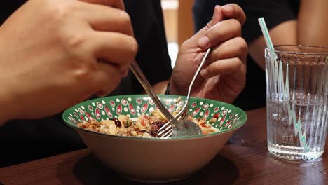 Close-up of hands using spoon and fork to eat rice meal in patterned bowl, with glass of water on wooden table, natural indoor lighting