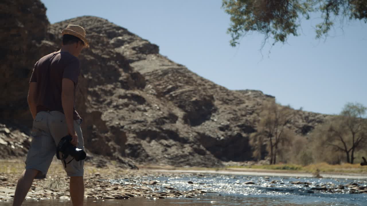 Handheld shot of a photographer tourist wearing a stylish hat carrying a camera walking through a riverbed in Africa with a mountain in the background.