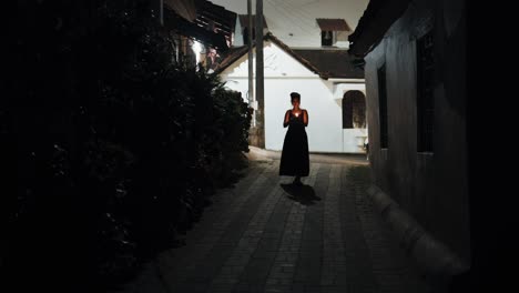 검은 드레스를 입은 예쁜 여성이 모자이크 바닥 위의 어두운 거리를 걷고 있는 모습, 개가 배경에서 그녀를 바라보는 동안 손에 불이 켜진 촛불을 들고 있는 모습