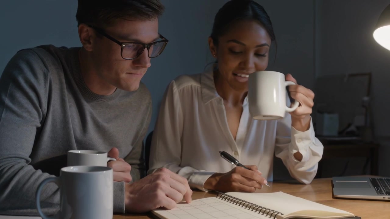 Pointing, man wearing sweater guiding woman wearing blouse writing in planner at desk with pen