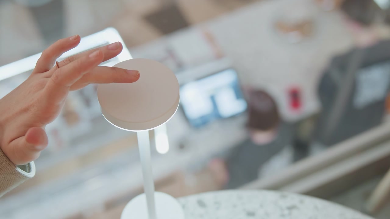 Primo piano di una mano sulla superficie superiore sensibile al tocco di un'elegante lampada da tavolo bianca che ne dimostra la funzionalità. Lo sfondo presenta un'illuminazione soffusa ed elementi sfocati che suggeriscono un ambiente rilassante.