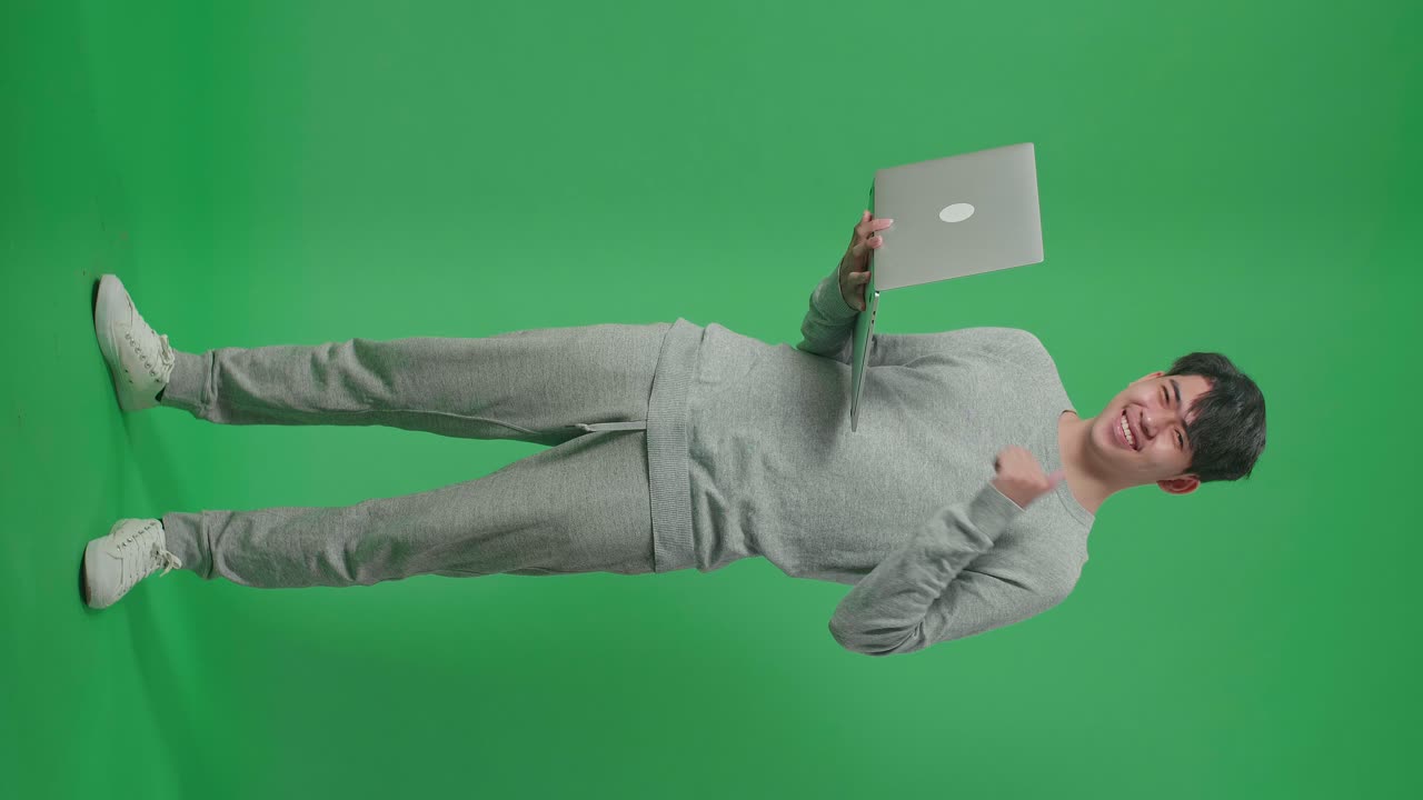 cuerpo lleno de hombre asiático sonriente mostrando pulgares hacia arriba gesto mientras escribe en la computadora en el estudio de pantalla verde