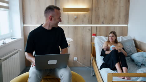 cheerful caucasian man looks at computer screen smiling. male turns to his wife sitting on the bed and breastfeeding a baby.