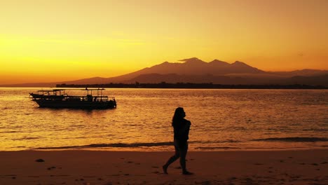 silhouette of girl walking along tranquil exotic beach at sunset with golden sky reflecting on calm lagoon full of boats in bali