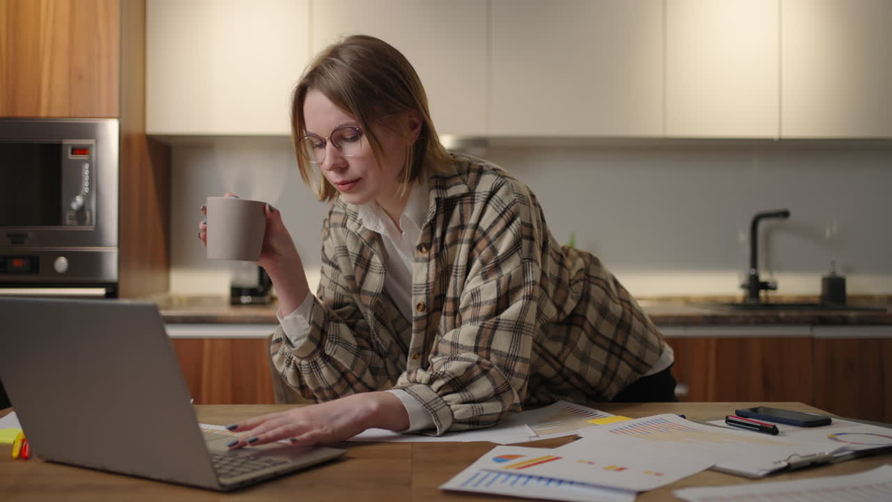 A young beautiful business woman works from home with a cup of coffee and looks into a laptop screen standing at the table. View mail and drink coffee. Remote work from home