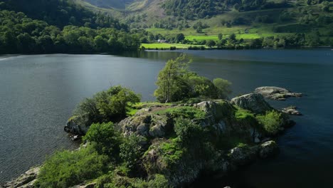 Flying-low-over-treetop-on-small-island-in-dark-lake-and-towards-lush-green-wooded-valley-with-reveal-of-mountains-beyond-on-bright-summer-day