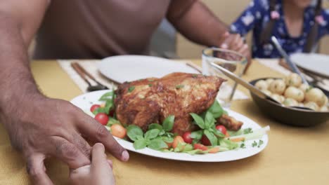 Happy-biracial-father,-son-and-daughter-saying-grace-at-dinner-table-in-garden,-slow-motion