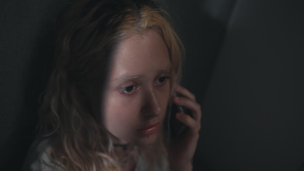 슬픈 젊은 금발 소녀가 어두운 회색 집의 회색 벽 근처에서 전화로 이야기