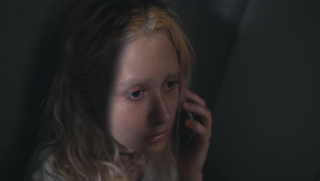 joven rubia triste hablando por teléfono cerca de una pared gris en una casa gris oscuro