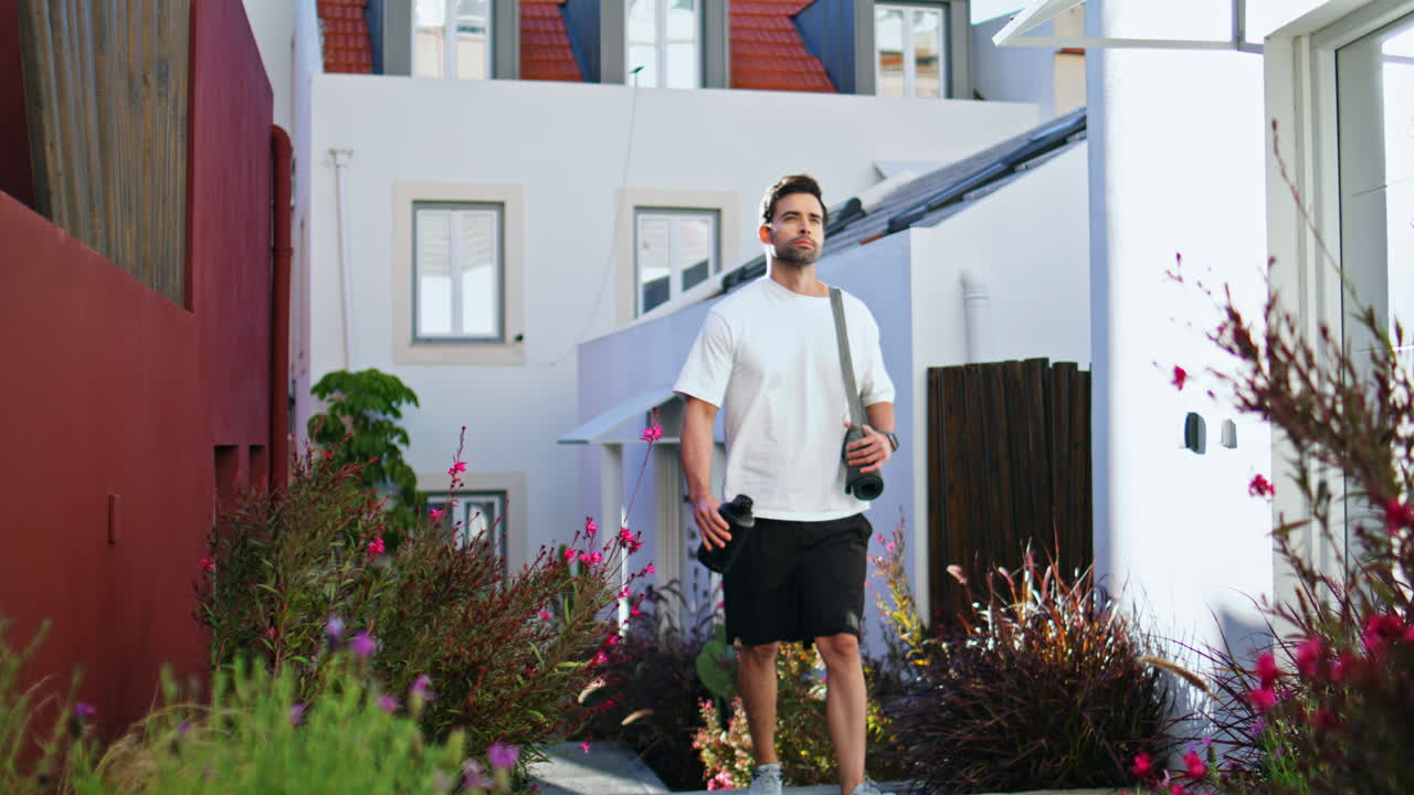 Man walking flowered street holding water bottle and sport mat. Handsome guy