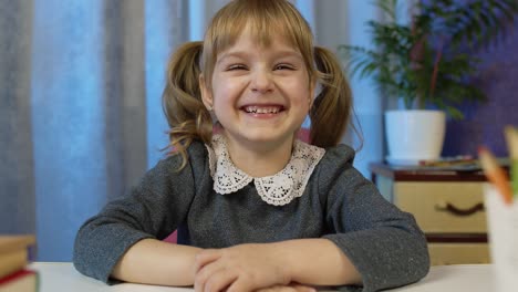 child girl pupil studying homework during online lessons at home, kid laughing, looking at camera