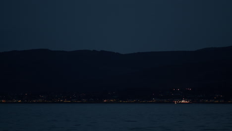 boat drives along coast with lights on vessel and on homes below mountains at the water