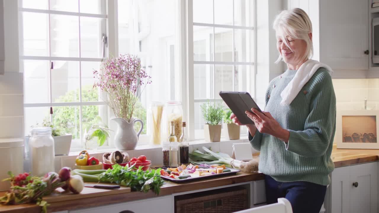mujer caucásica mayor feliz de pie en la cocina y preparando la cena, usando una tableta