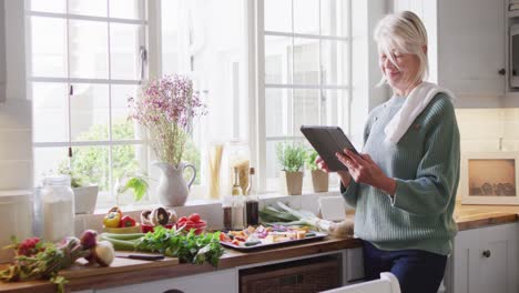 happy senior caucasian woman standing in kitchen and preparing dinner, using tablet
