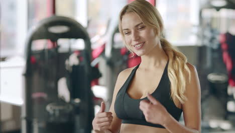 Fitness-woman-showing-thumb-up-and-winking-eyes-on-sport-club-background