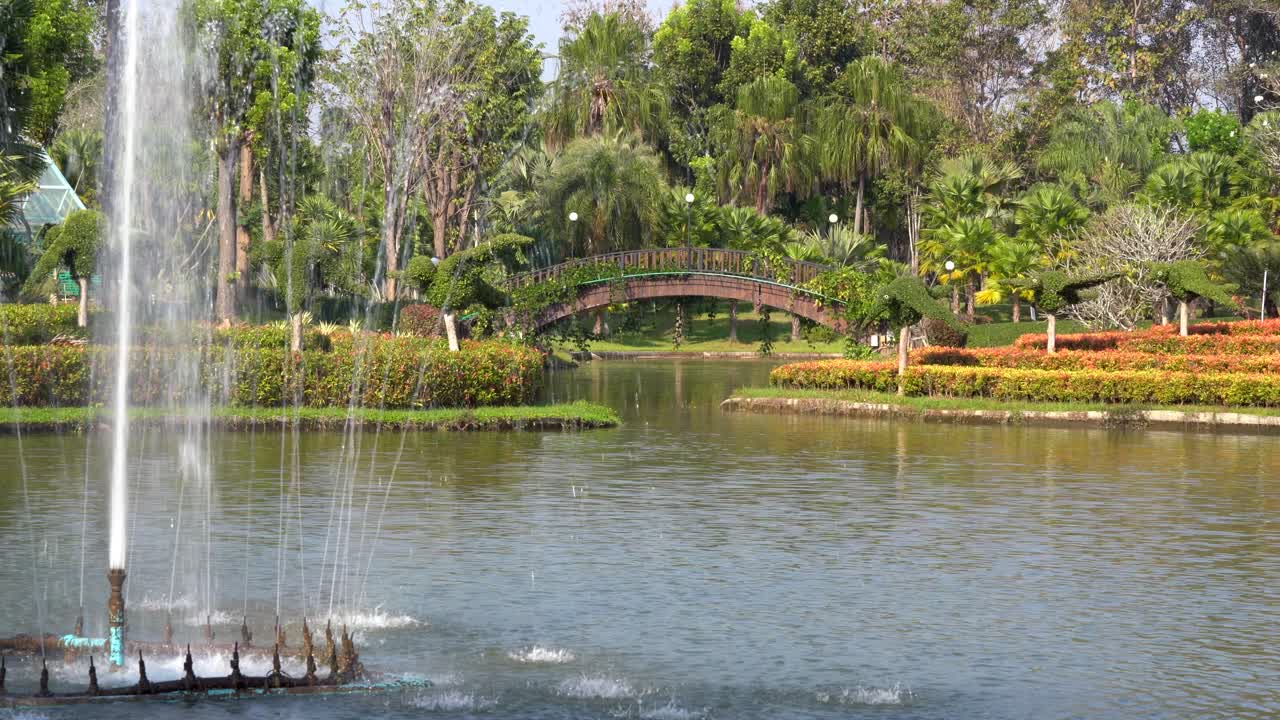 una fuente rociando agua en un estanque con un puente arqueado en el fondo rodeado de hermosas plantas
