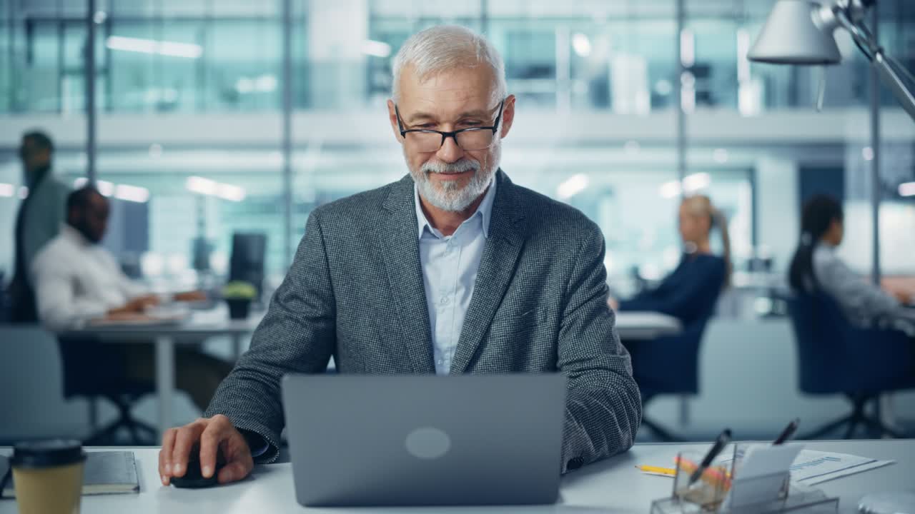 Modern Office: Portrait of Successful Middle Aged Bearded Businessman Working on a Laptop at his Desk. Smiling Corporate Worker. Multi-Ethnic Workplace with Happy Professionals. Front View Static Shot