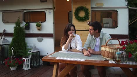Portrait-of-cute-european-couple-man-and-woman-drinking-white-wine-while-sitting-together-at-wooden-table-outdoors.-Stylish,-static-wheels-house-on-background
