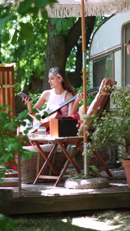 woman playing guitar and looking at a smartphone on a deck outside a campervan