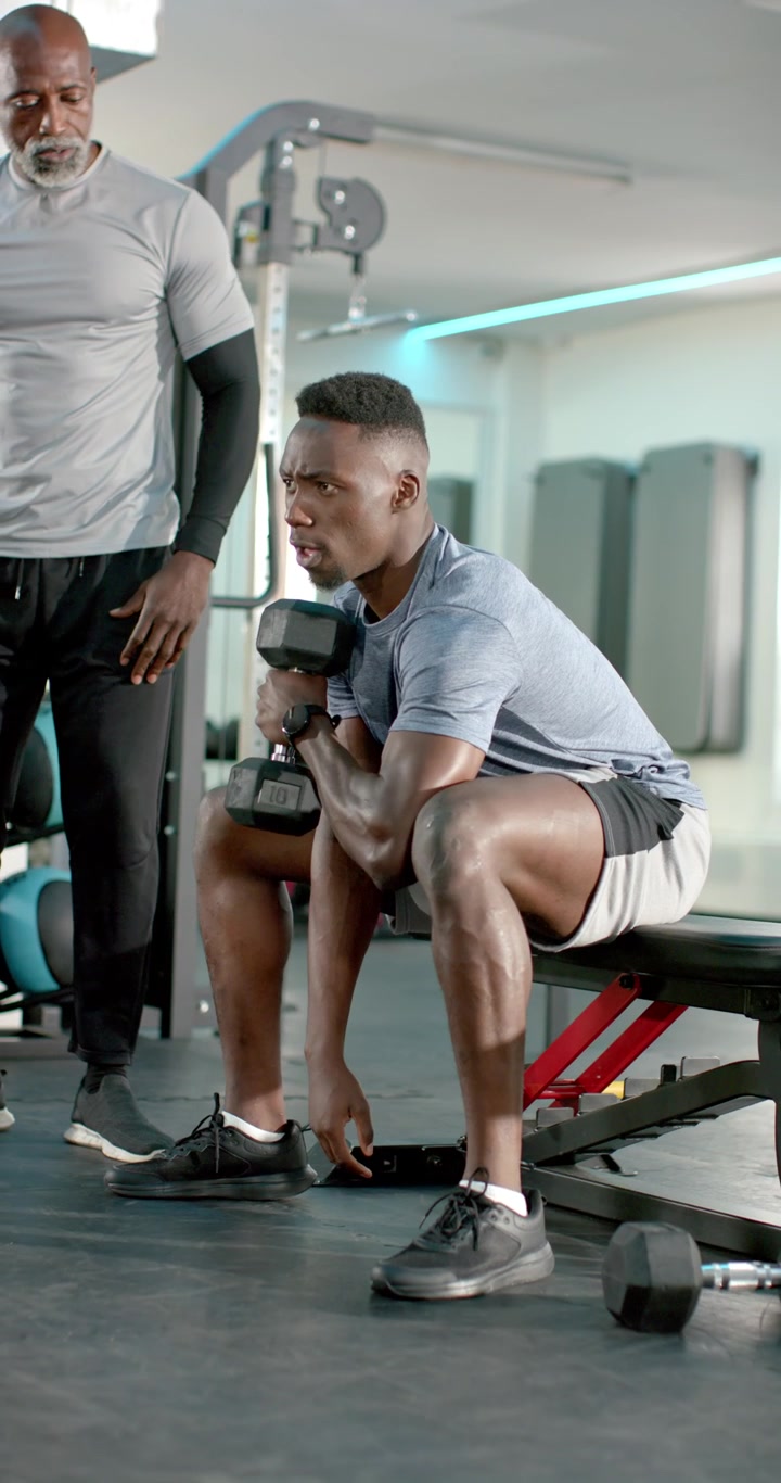 Vertical video: African American man curling dumbbell at bench while trainer guiding form in gym
