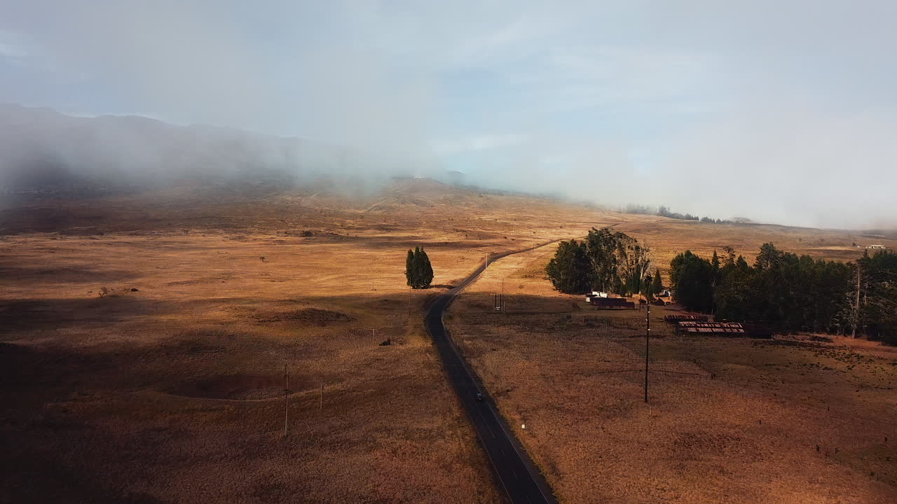 Moving aerial shot of a car driving on a highway through golden fields on the Mauna Kea hillside. Mist on the horizon. Cinematic 4k with an orange and teal look.