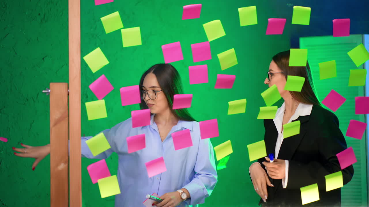 Ladies wearing glasses stand behind the glass board covered with stickers. Women disagree or arguing about work issues.