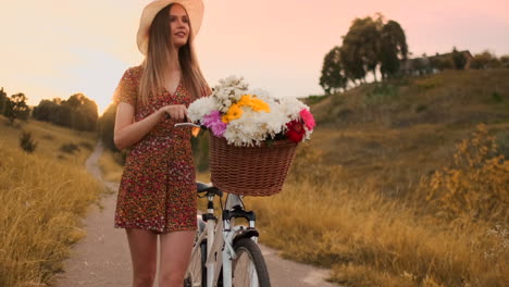 Destello-De-Lente:-Mujer-Feliz-Y-Sonriente-Con-Vestido-Corto-Anda-En-Bicicleta-Con-Una-Canasta-Y-Flores-En-El-Parque-Con-árboles-Verdes-Alrededor-Durante-El-Amanecer.-Toma-En-Cámara-Lenta