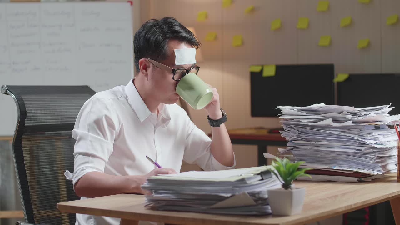 병든 아시아인 남자가 사무실에서 문서와 함께 열심히 일하면서 물을 마시고 있습니다.