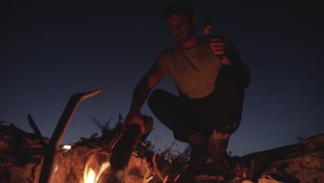 caucasian man moving dead wood log in firepit on bright clear evening sky with half moon in distance, low angle close up pan