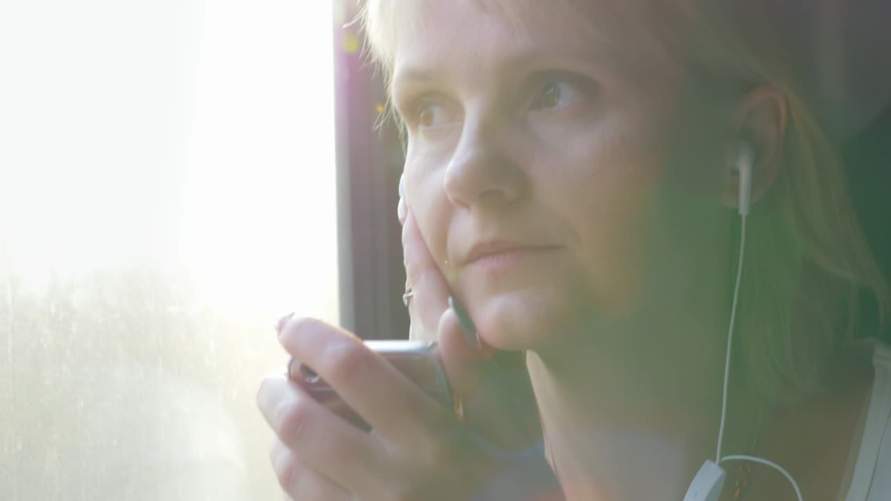 молодая белая женщина едет в купе поезда, держит смартфон в руке, слушает музыку на наушниках