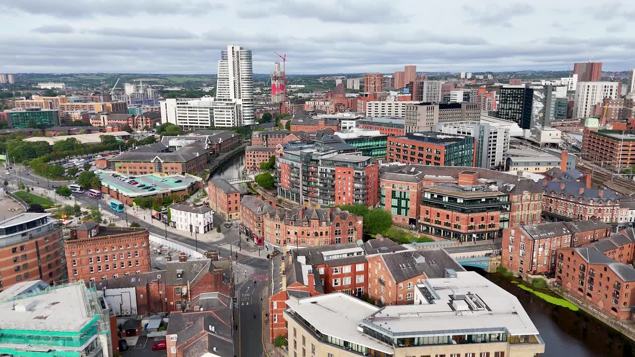 Kamera drona płynnie panoramuje nad centrum Leeds, ukazując wiktoriańskie budynki, nowoczesne wieże i rzekę Aire w jasnym świetle dziennym z delikatnym zachmurzeniem.