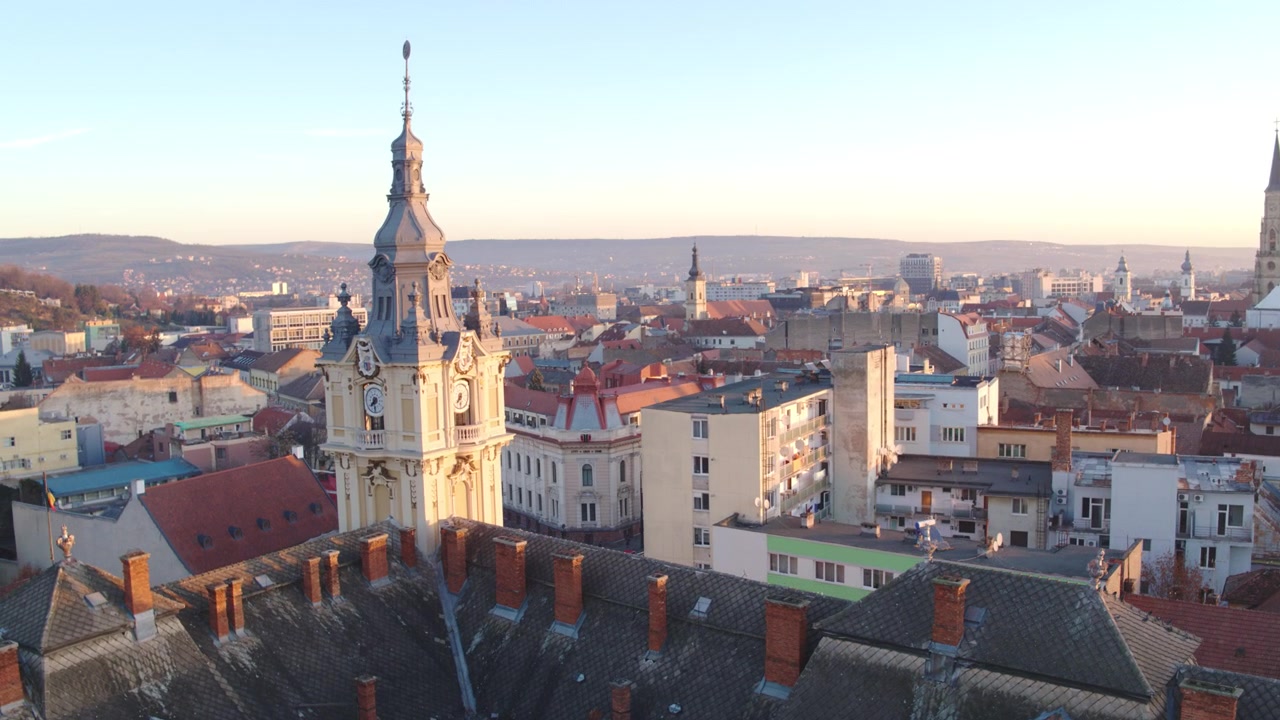 Ripresa aerea che si eleva sopra vecchi tetti di tegole per rivelare la storica torre dell'orologio del Municipio e lo skyline della città di Cluj-Napoca sotto un cielo mattutino limpido e blu.